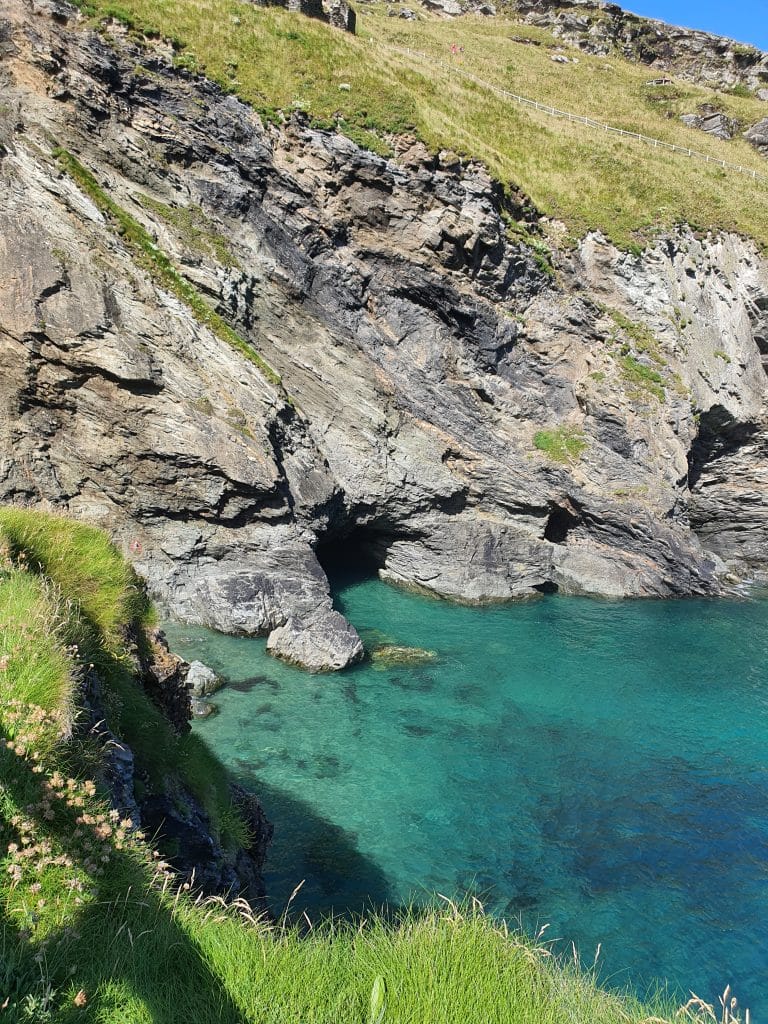 Sover Travel - A rocky cliffside with patches of green grass overlooks the vibrant turquoise waters near Merlins Cave in Cornwall. The clear water reveals rocks beneath the surface, all bathed in bright sunlight under a pristine blue sky.