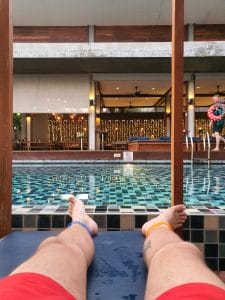 Sover Travel - Relaxing poolside on a blue lounge chair, legs extend in red swim trunks adorned with colorful anklets. The backdrop evokes a Cornish charm reminiscent of Merlins Cave, featuring a deck with chairs, tables, and string lights under a cozy covered area.