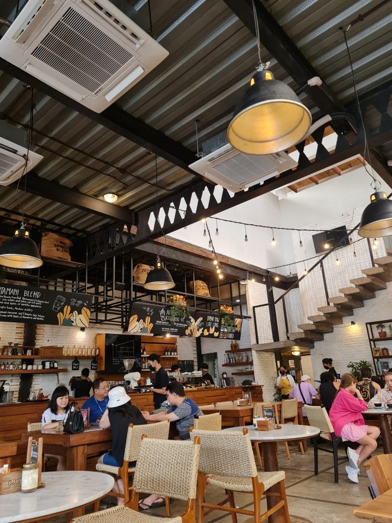 Sover Travel - People sitting and chatting inside a cozy café with wooden tables and chairs. The café features industrial-style lighting, a staircase, and a blackboard menu. Several potted plants adorn the shelves above the counter.