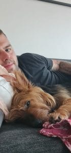 Sover Travel - A person is lying on a couch next to a small brown dog with long fur. The dog is resting its head on a pink pillow, and their eyes are partly closed. The person is wearing a black shirt, and part of a tattoo is visible on their arm.