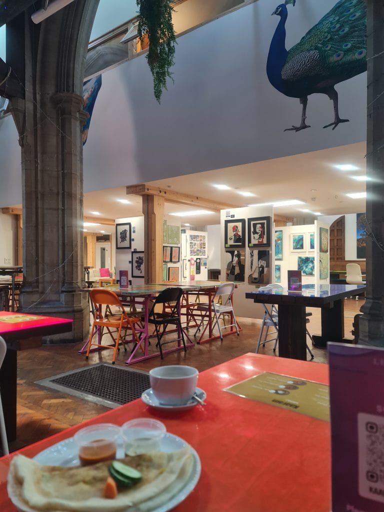 Sover Travel - Art gallery interior with colorful folding chairs around tables. A peacock mural is visible on the wall. Foreground shows a table with a plate of flatbread, small sauce cups, and a coffee cup. Bright lights and artworks are displayed on the walls.