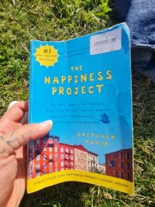 Sover Travel - Close-up of a hand holding a worn copy of The Happiness Project by Gretchen Rubin. The blue cover features the title and colorful buildings beneath. The book is placed on grass, and a persons foot is partially visible in the background.