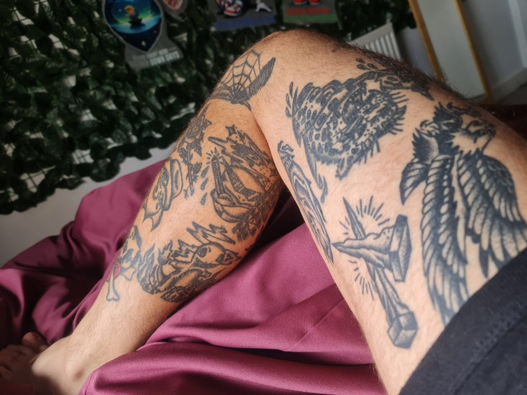 Sover Travel - A persons tattooed legs are stretched out on a purple bedspread in their Bristol apartment. The tattoos, showcasing a spider web, animals, and abstract designs, tell unique stories. In the background, a dark green leafy curtain complements framed pictures lining the wall.