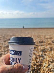 Sover Travel - Close-up of a hand holding a Greggs coffee cup on a pebble beach. Blurred background shows the sea under a partly cloudy sky.