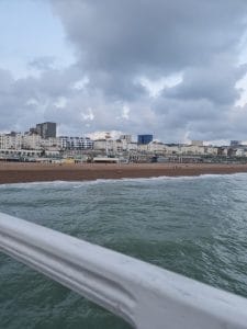 Sover Travel - A coastal city view features a pebble beach along Kennedy Street with a row of diverse buildings and a cloudy sky. People stroll by Recovery Hub on the beach, and the foreground shows a railing above the greenish sea.