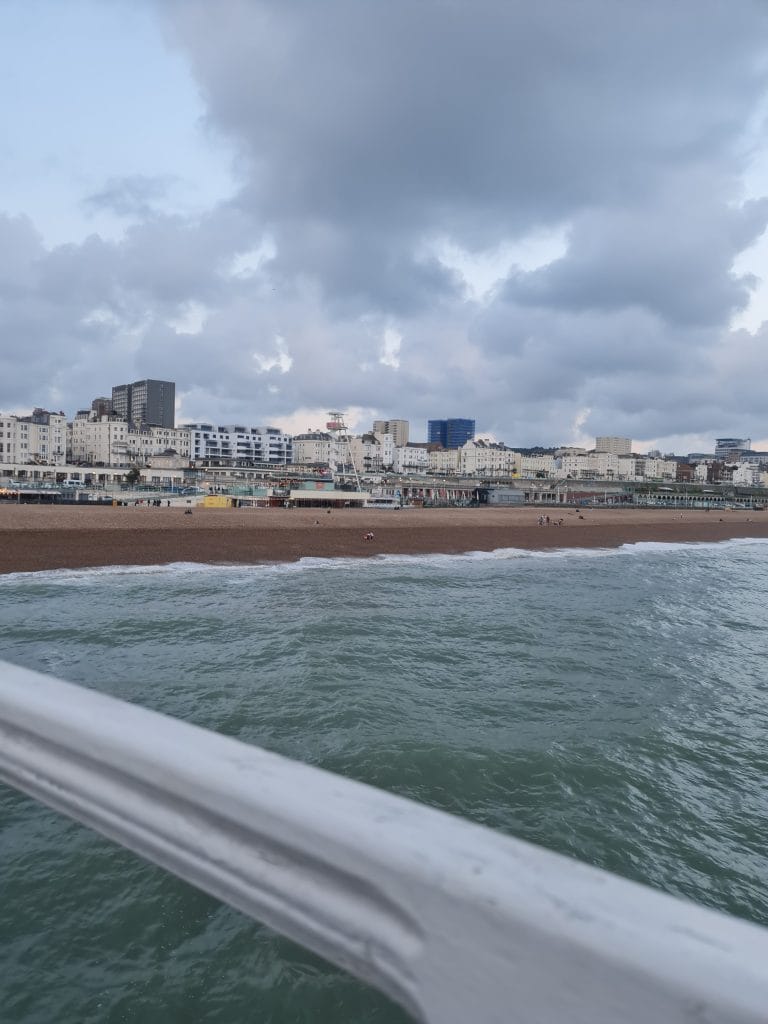 Sover Travel - A coastal city view features a pebble beach along Kennedy Street with a row of diverse buildings and a cloudy sky. People stroll by Recovery Hub on the beach, and the foreground shows a railing above the greenish sea.