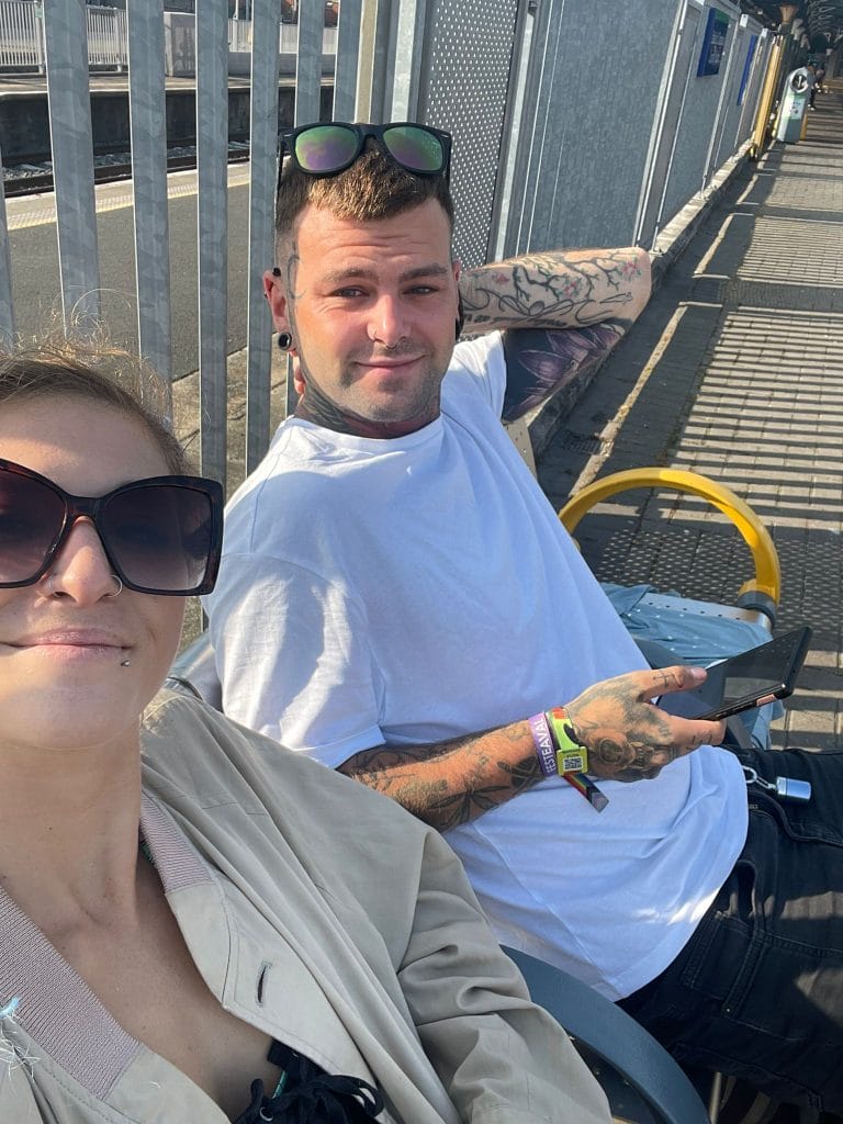 Sover Travel - Two people sitting on a bench at a train station. The person on the left wears large sunglasses and a beige jacket, and the person on the right has tattoos, a white shirt, and sunglasses on their head while holding a phone. Rails and platform visible.