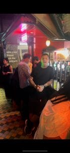 Sover Travel - A man stands at a lively bar, wearing a black shirt and shorts, with tattoos on his arms. He is smiling and holding onto the counter. The bar has colorful lights and a checkered floor, with several people in the background.