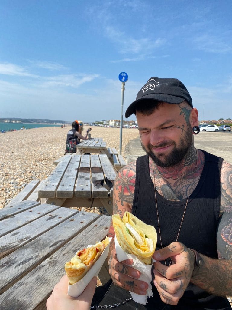 Sover Travel - A tattooed man exudes presence in a black tank top and cap, smiling as he holds a wrap on the beach. Another hand offers him a second wrap. Wooden benches and a cyclist create dynamic energy in the background, set against the blue sky and pebbled shore.