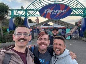 Sover Travel - Four men are smiling and posing for a selfie in front of the entrance to Thorpe Park, where a large sign displays the parks name. Two of the men, one showcasing a vibrant tattoo, are holding their arms up in excitement. Its a sunny day, perfect for an adventure before heading back to Bristol.