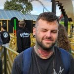 Sover Travel - A man with a Stush Tattoo and a backpack stands in an amusement park queue in Bristol, squinting slightly with one eye closed. People wait patiently behind him. The atmosphere is casual and relaxed.