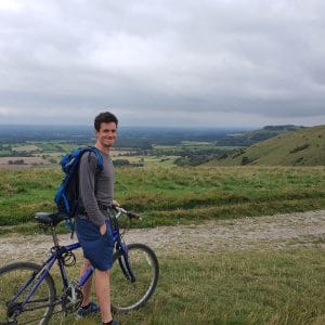 Sover Travel - A person with a backpack stands beside a bicycle on a grassy hilltop, smiling. The background features a sprawling landscape with fields, trees, and a cloudy sky.