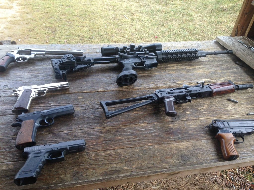 Sover Travel - A wooden table outdoors displaying an assortment of firearms, including rifles and pistols. The background shows grass and part of a wooden structure.