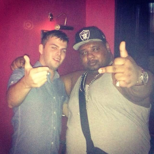 Sover Travel - Two men pose in a dimly lit room with red walls. The man on the left wears a blue shirt and points towards the camera. The man on the right is in a gray tank top and hat, also pointing at the camera. Both appear to be in a casual setting.