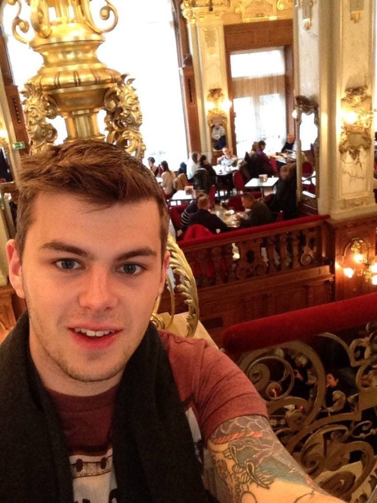 Sover Travel - A person with tattoos takes a selfie in an elegant, ornate restaurant featuring golden decorations and chandeliers. Several diners can be seen in the background sitting at tables.
