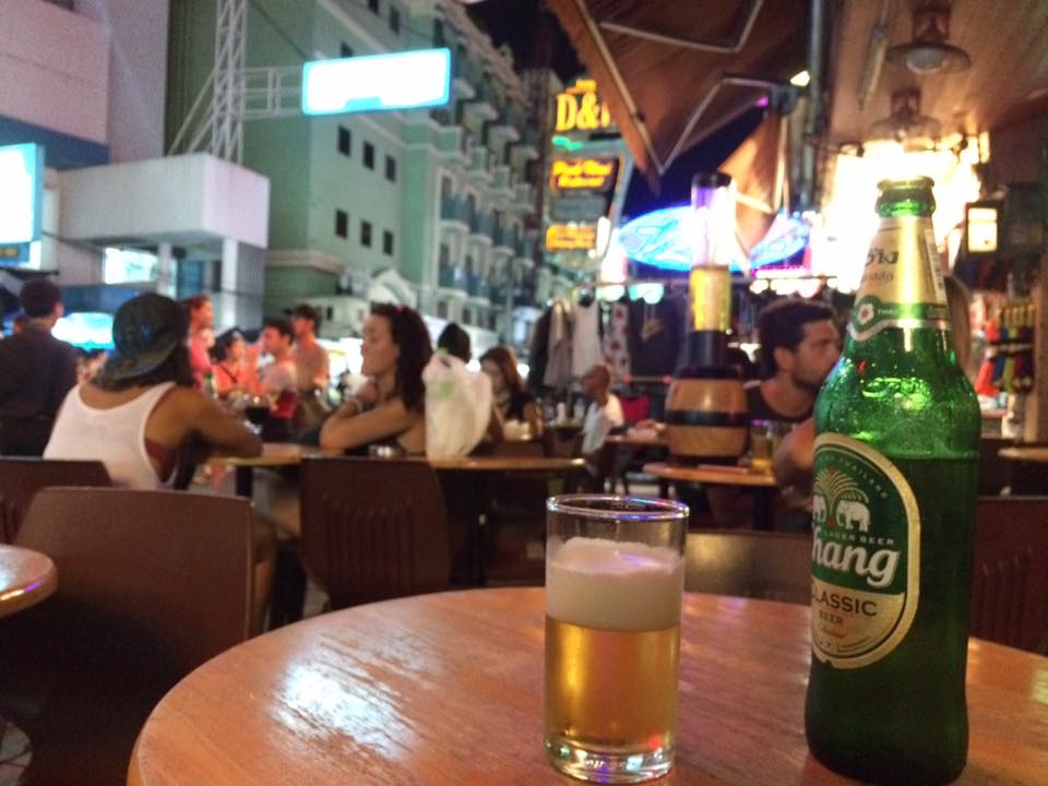 Sover Travel - A bustling outdoor café scene at night with people sitting at tables. A Chang beer bottle and a half-filled glass are on a wooden table in the foreground. Bright neon signs illuminate the background, creating a lively atmosphere.
