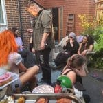 Sover Travel - A group of people gathered outdoors at a casual gathering, seated and standing around a patio. Food and beverages are on a table in the foreground. The setting is relaxed, with brick walls and plants in the background.