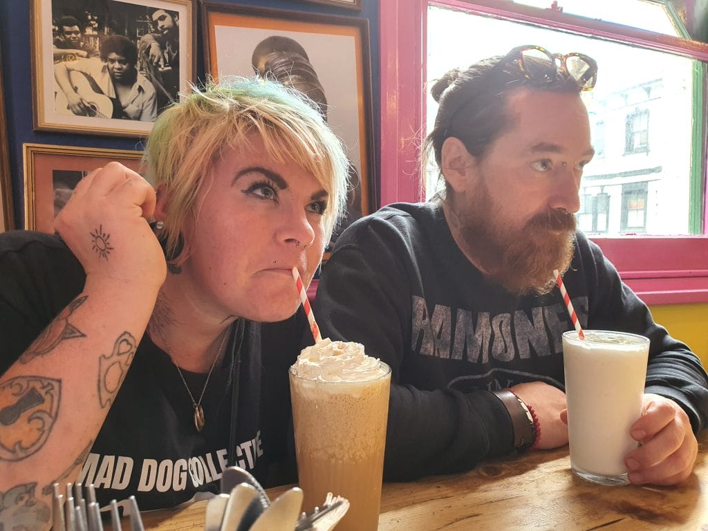 Sover Travel - Two people sitting at a table, each with a drink. The person on the left has colorful hair and tattoos, sipping from a whipped cream-topped beverage. The person on the right, with a beard and sunglasses on their head, holds a milkshake. Theyre indoors with framed photos on the wall behind them.