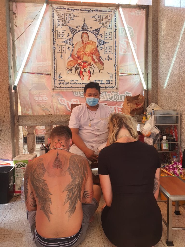 Sover Travel - A tattoo artist in a mask sits in a small studio with two clients in front of him. The man has wing tattoos on his back; the woman has long blonde hair. A poster on the wall features a monk and Thai script. Supplies are visible around the studio.