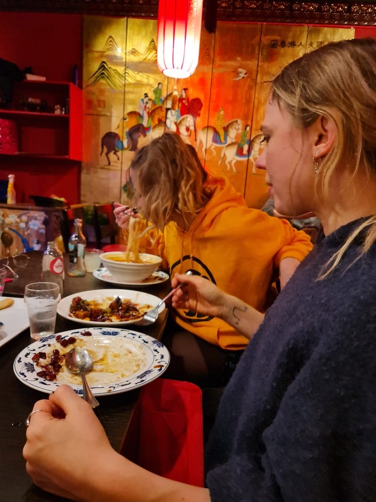 Sover Travel - Two people are seated at a restaurant table, eating from plates of food. The person in the foreground holds a spoon and wears a dark top. The person in the background, wearing a yellow hoodie, is using chopsticks. The decor features a vibrant mural and red lighting.