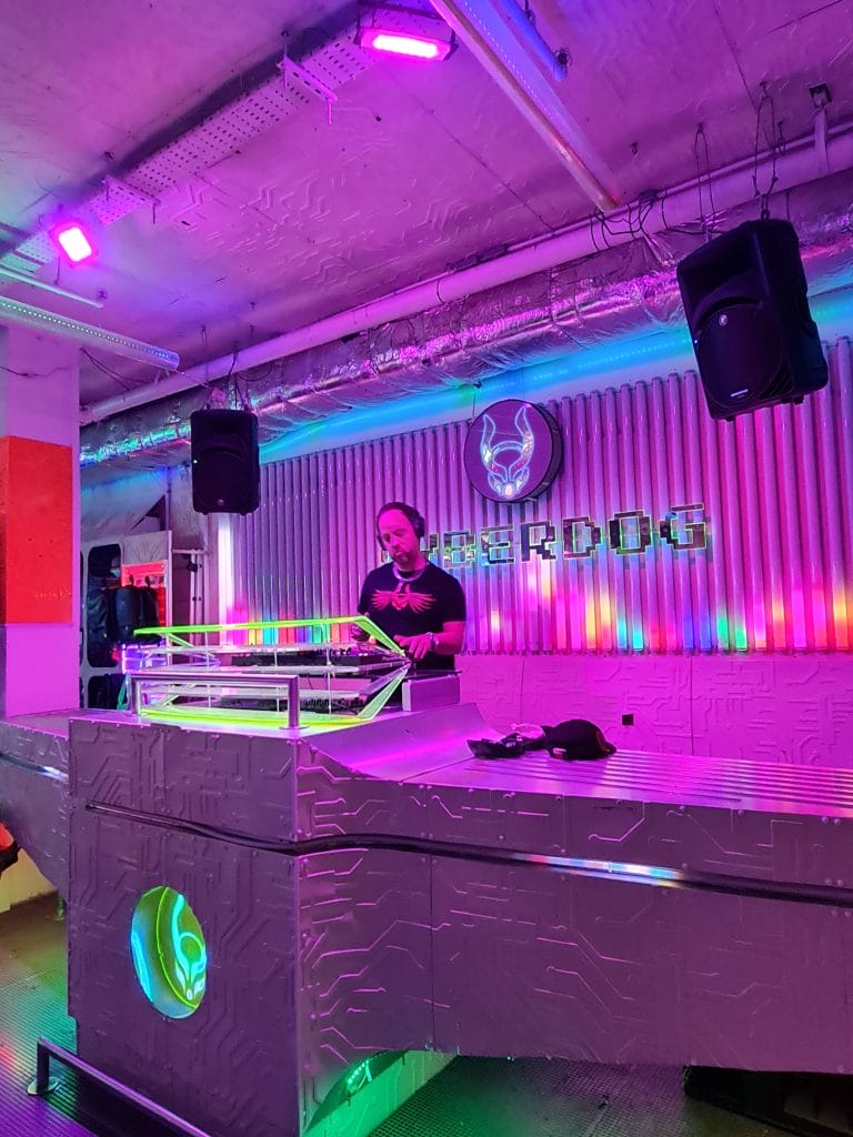 Sover Travel - A DJ stands behind a futuristic, illuminated DJ booth in a club with vivid neon lighting. The background features corrugated metal walls and a logo. Speakers are mounted on the walls, and colorful lights create a vibrant atmosphere.