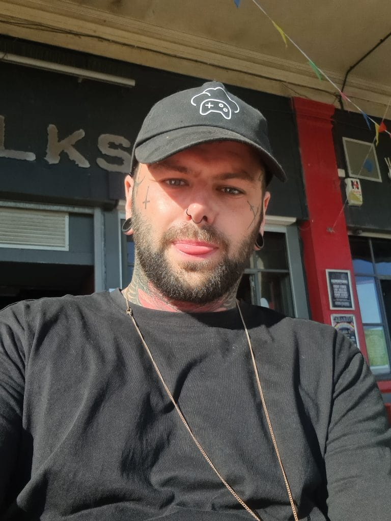 Sover Travel - A bearded person wearing a black cap and shirt sits outside, with tattoos on their face and neck. Bright sunlight casts shadows, and colorful flags hang in the background, attached to a building with red and black trim.