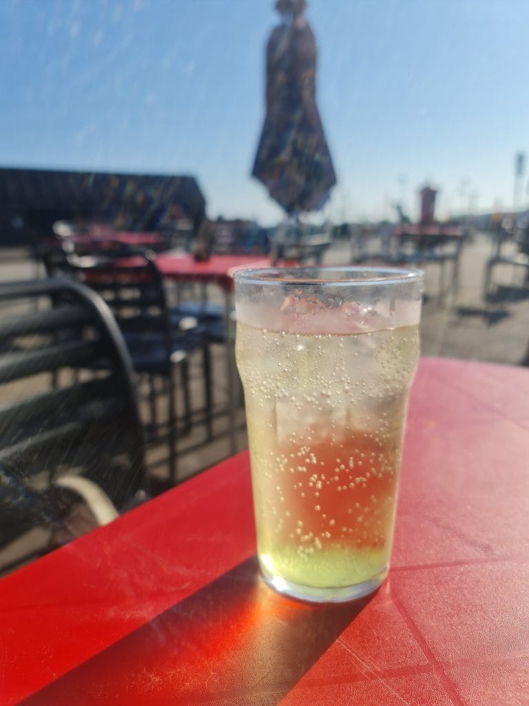 Sover Travel - A glass of sparkling beverage with ice sits on a red table outdoors. The background is a blurred figure of a person walking and several empty chairs and tables under a clear blue sky.