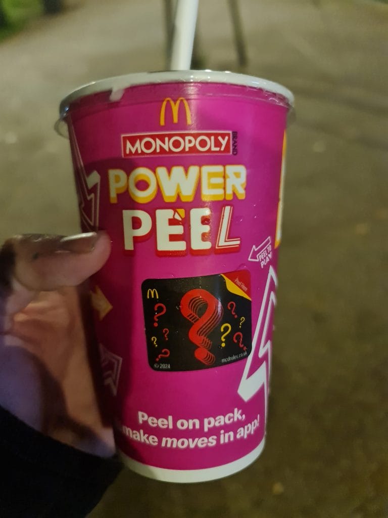 Sover Travel - A hand holding a red McDonalds Monopoly cup with the words Power Peel printed on it. There is a game sticker with question marks on the cup. A white straw is inserted in the drink. The background is blurred.