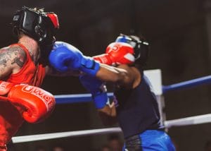 Sover Travel - Two boxers wearing protective headgear engage in a match. One boxer in red gloves delivers a punch, while the other in blue gloves attempts to block. They are in a boxing ring with blurred background, emphasizing the action.