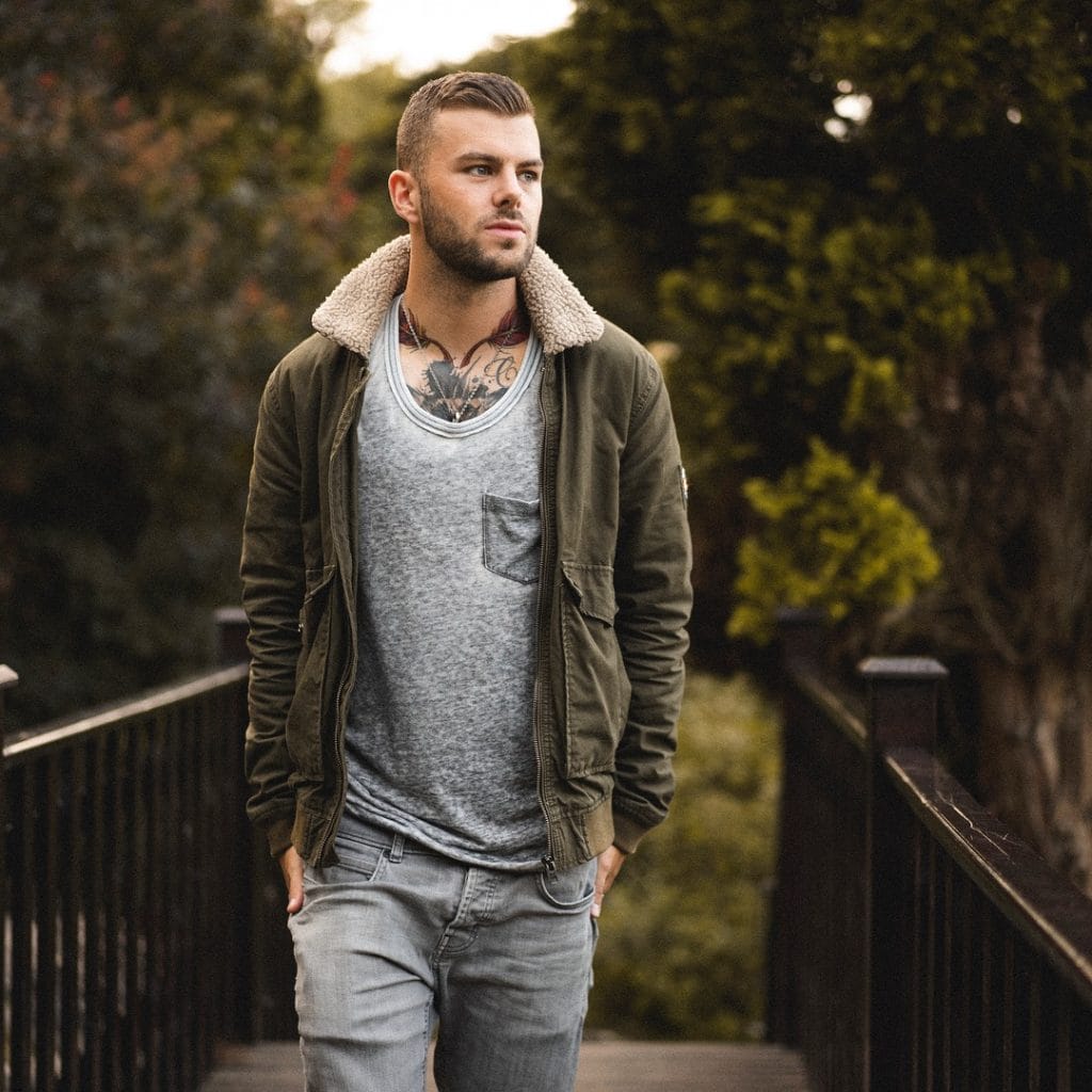 Sover Travel - A man with short hair and tattoos is walking on a wooden bridge. Hes wearing a green jacket with a fur collar, a gray shirt, and jeans. The background features blurred trees and foliage.
