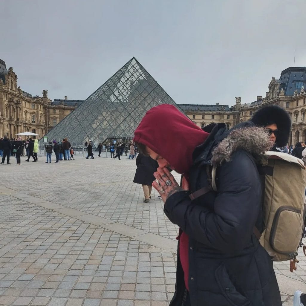 Sover Travel - A person in a red hood and black coat is standing with hands clasped, near the glass pyramid entrance of the Louvre Museum. Another person in a dark jacket and sunglasses is nearby. Crowds are scattered around on a cloudy day.