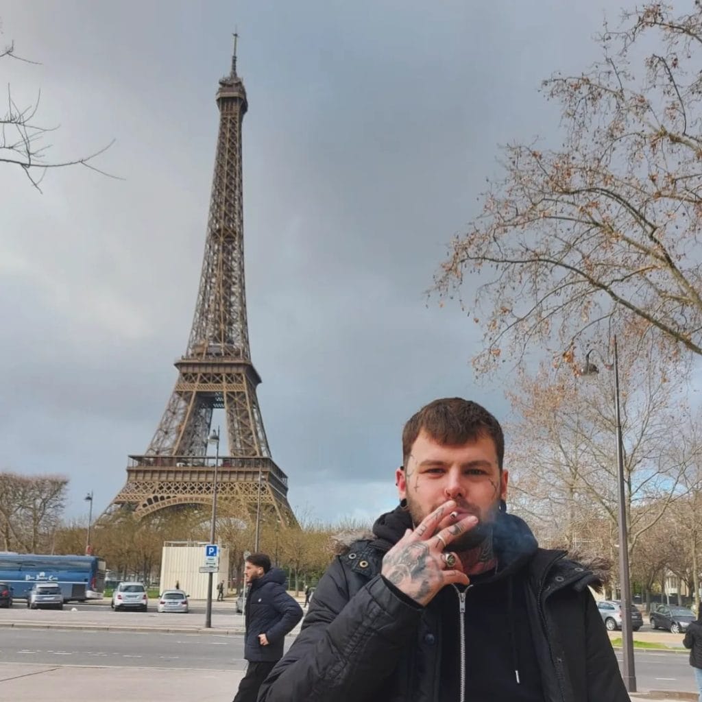 Sover Travel - A person with facial hair and tattoos smokes a cigarette in the foreground. The Eiffel Tower is prominently visible in the background. The scene includes winter trees and cloudy skies, with parked vehicles in the distance.