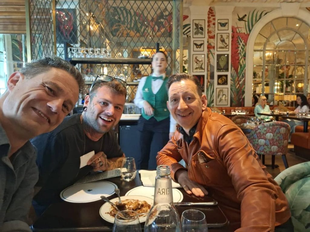 Sover Travel - Three people smile at a table in a restaurant with a server walking behind them. The table has plates and a water bottle. The restaurant decor is ornate with mirrors and art on the walls.