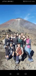 Sover Travel - Group of ten people posing together on rocky terrain with a large, dormant volcano in the background under a clear blue sky. Some wear backpacks, and most are dressed in casual outdoor attire.