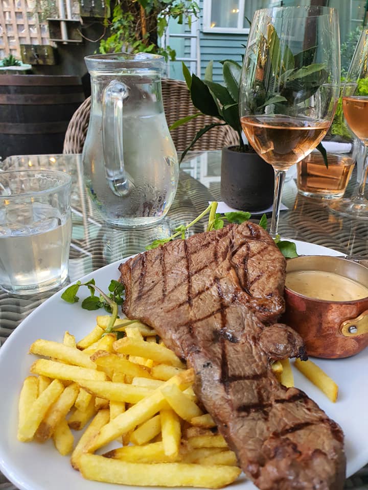 Sover Travel - A plate with a grilled steak and fries is on a table, accompanied by a small copper pot of sauce. There are two glasses of rosé wine, a jug of water, and a glass of water nearby. Plants and wicker furniture are in the background.
