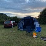 Sover Travel - A blue-domed tent is set up in a grassy field next to a red car. Two blue camping chairs are nearby, with a yellow backpack on the ground. The sky is partly cloudy, and its either sunrise or sunset. Trees and bushes are in the background.