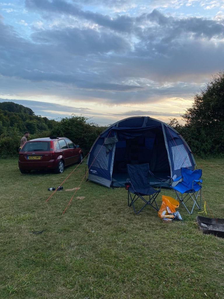 Sover Travel - A blue-domed tent is set up in a grassy field next to a red car. Two blue camping chairs are nearby, with a yellow backpack on the ground. The sky is partly cloudy, and its either sunrise or sunset. Trees and bushes are in the background.