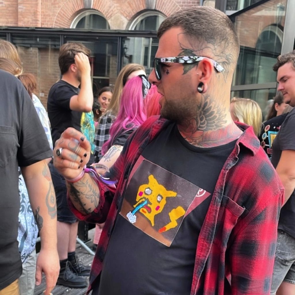 Sover Travel - A person wearing a red flannel shirt and a colorful graphic t-shirt featuring a cartoon character stands in a crowd of people outdoors. The person has tattoos, large earrings, and is wearing sunglasses. Buildings are visible in the background.
