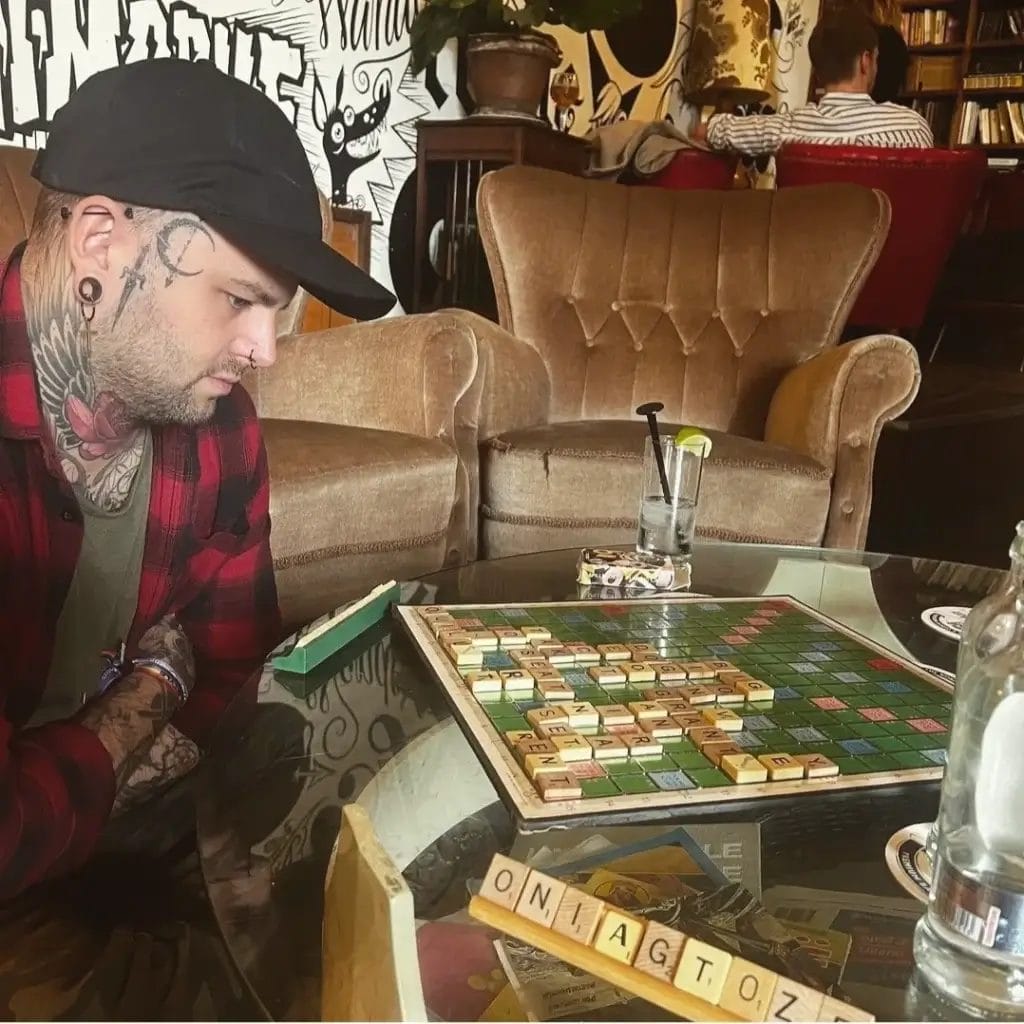 Sover Travel - A person wearing a black cap and plaid shirt plays Scrabble, focusing on the board. The room has cozy armchairs, a mural on the wall, and another person reading in the background. A drink with a lime wedge is on the table.