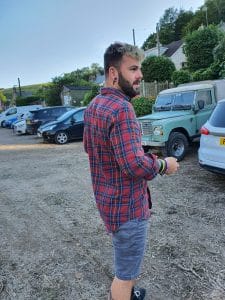 Sover Travel - A person wearing a red and blue plaid shirt and gray shorts stands outdoors near a row of parked cars. Trees and hills are visible in the background, and the person is looking to the right.