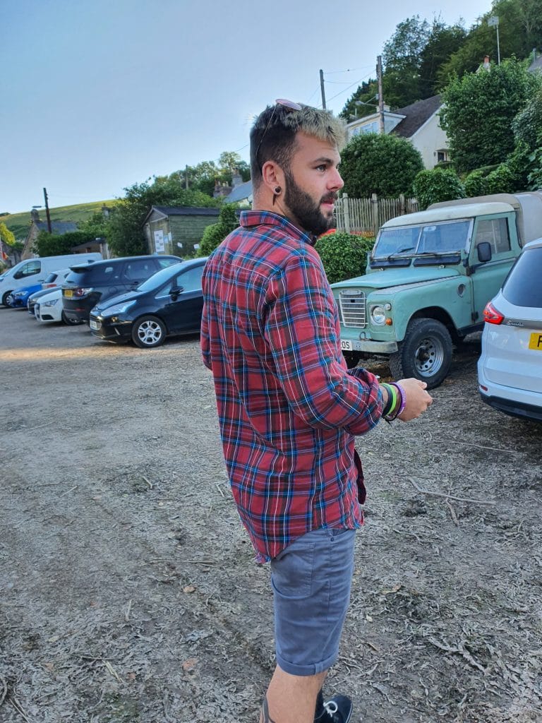 Sover Travel - A person wearing a red and blue plaid shirt and gray shorts stands outdoors near a row of parked cars. Trees and hills are visible in the background, and the person is looking to the right.