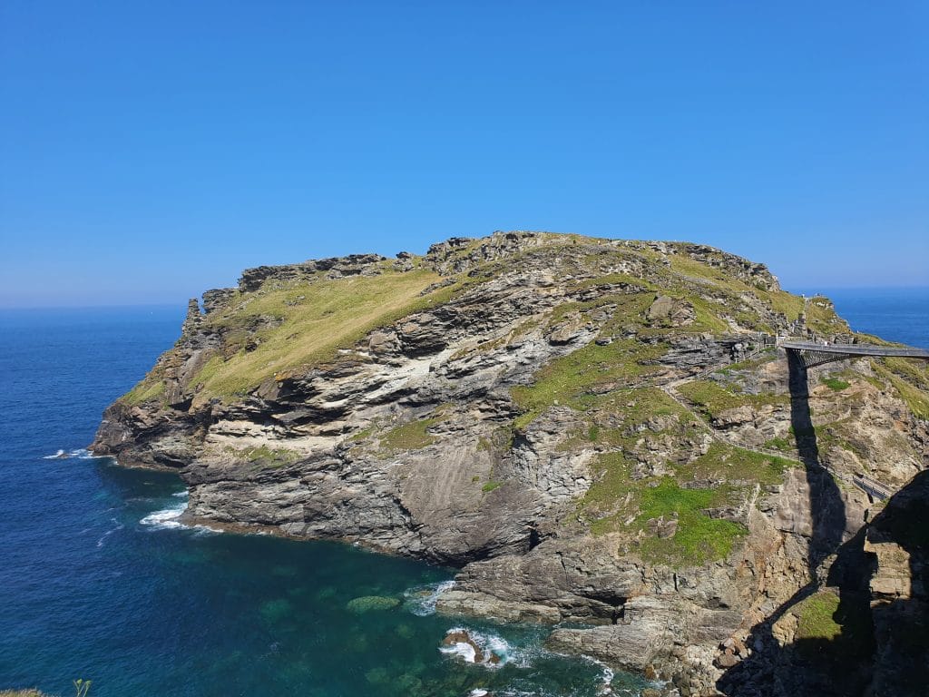 Sover Travel - A rocky cliff extends into the bright blue sea under a cloudless sky. A narrow bridge connects the land to the cliff, with small, distant figures walking on it. The cliff is covered with patches of grass and rugged rock formations.