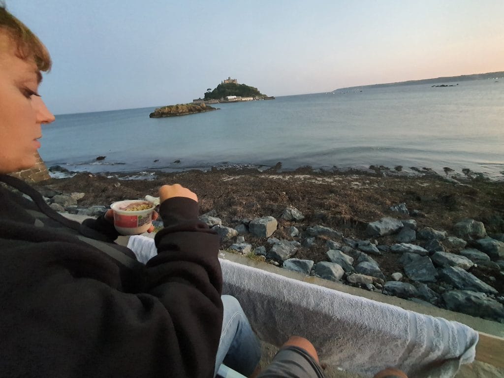 Sover Travel - A person sitting on a rocky shoreline, eating from a small container. The ocean is in the background with an island featuring a castle-like structure. The sky is clear, suggesting its either dawn or dusk. The person is wearing a dark jacket.