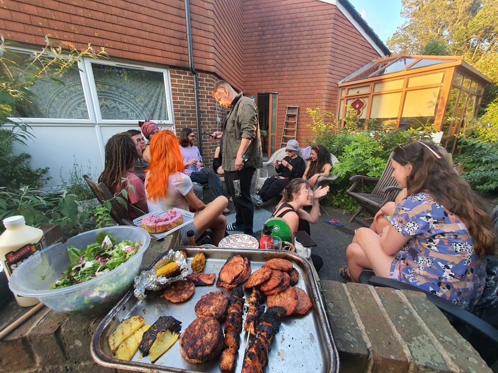 Sover Travel - A group of people enjoying a backyard gathering. Grilled food and salad are on the table in the foreground. Various individuals are sitting and talking, with some engaging each other lively. The setting has plants and a brick building in the background.