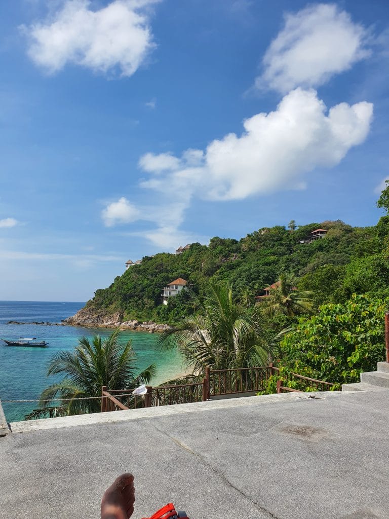 Sover Travel - View of a coastal landscape in Koh Tao, Thailand, with a clear blue sea, lush green hills, and scattered houses. A small boat floats in the water. Palm trees and steps are in the foreground under a partly cloudy sky—a hidden beauty captured with just a glimpse of someones foot at the edge.