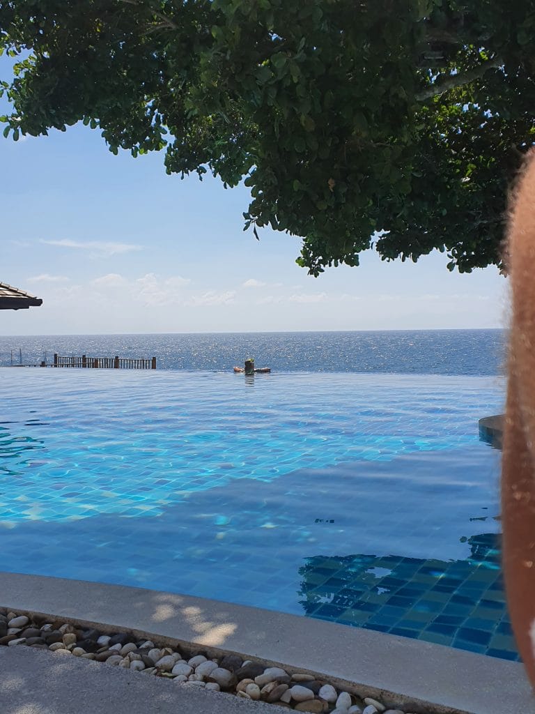 Sover Travel - Infinity pool overlooking a vast ocean under clear blue skies on the hidden beauty of Koh Tao. A person relaxes on a float, partially shaded by a large tree, with the rocky edge in the foreground.