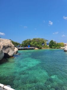 Sover Travel - The hidden beauty of Koh Tao reveals itself in the clear turquoise water, where large rocks rest in the foreground, surrounded by lush green trees under a bright blue sky with scattered clouds. A small dock stretches from the land into this tranquil Thai paradise.