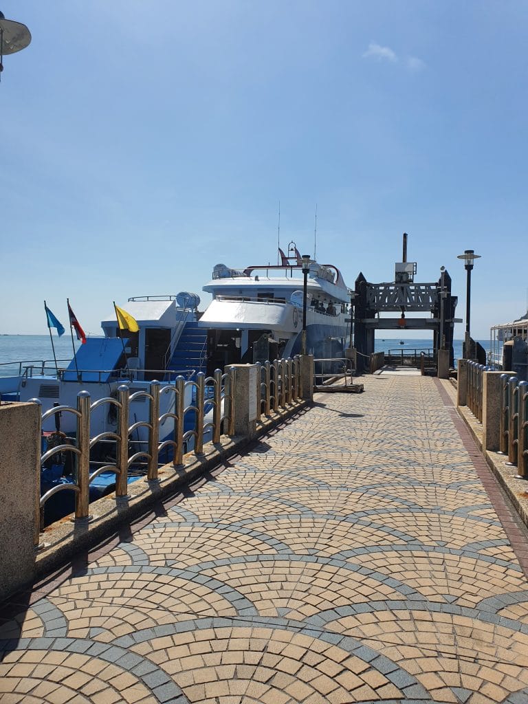 Sover Travel - A white yacht docked at a pier on a sunny day in Koh Tao, Thailand. The walkway is paved with a geometric pattern and bordered by a metal railing. Flags flutter on the left side of this hidden beauty, under the clear blue sky.