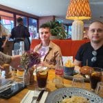 Sover Travel - Two people sit at a restaurant table on Koh Tao with various dishes, including a burger, fries, salads, and drinks. A hand extends over the table reaching for a plate. Theyre surrounded by a cozy setting with a decorative lamp and tropical plants reminiscent of Thailands charm.