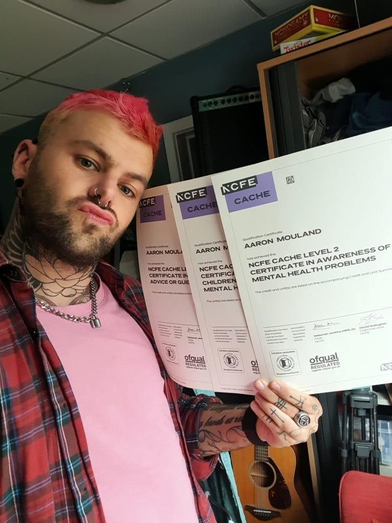 Sover Travel - In a sunlit room filled with musical equipment, a person with pink hair and tattoos confidently holds three certificates. Wearing a pink and plaid shirt, they exude confidence as they stand like an artist ready to perform on Maltas vibrant streets.
